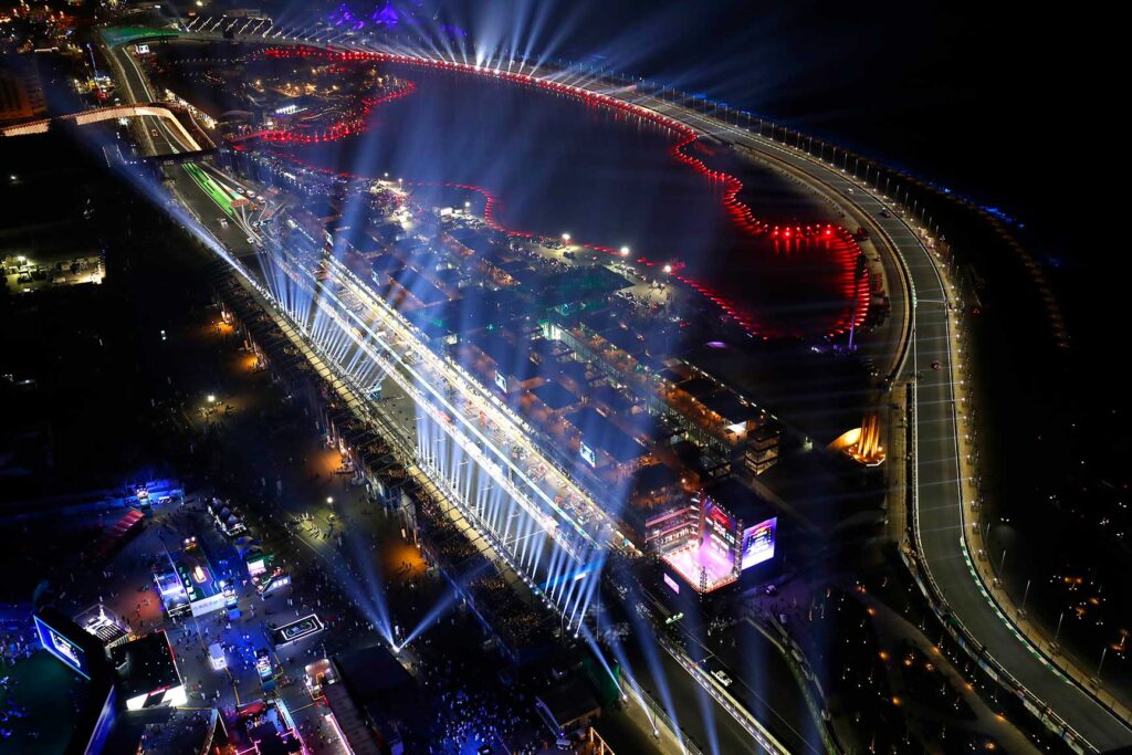 這張照片以高空視角拍攝沙烏地阿拉伯吉達街道賽道（Jeddah Corniche Circuit）的夜間景色。主看台與維修區被強烈的白色燈光打亮，整條直道光束向天空散射，營造出濃厚的夜賽氛圍。右側賽道沿著海岸彎曲延伸，紅色燈光在水岸邊形成波浪狀線條，點亮整片海濱。畫面下方為活動區與觀眾區，充滿展演燈光與人群活動，呈現 F1 週末熱鬧且科技感十足的場面。